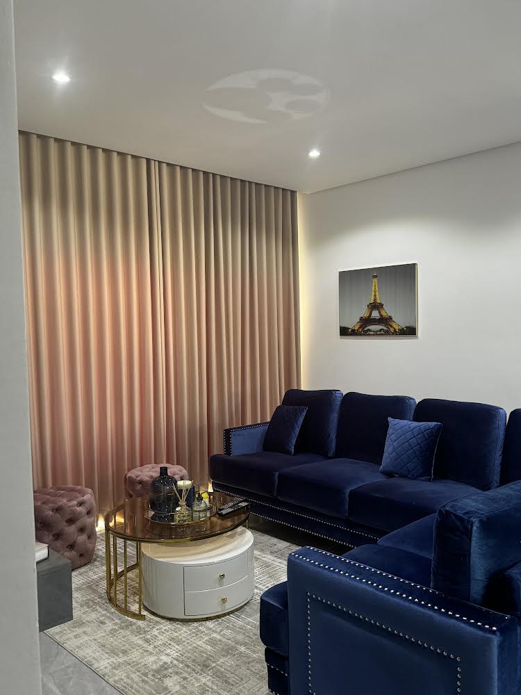 A lounge area features deep blue velvet sofas paired with tufted ottomans. A round coffee table sits at the center, adorned with decorative items. Light filters through vertical curtains, while a framed image of the Eiffel Tower hangs on the wall.