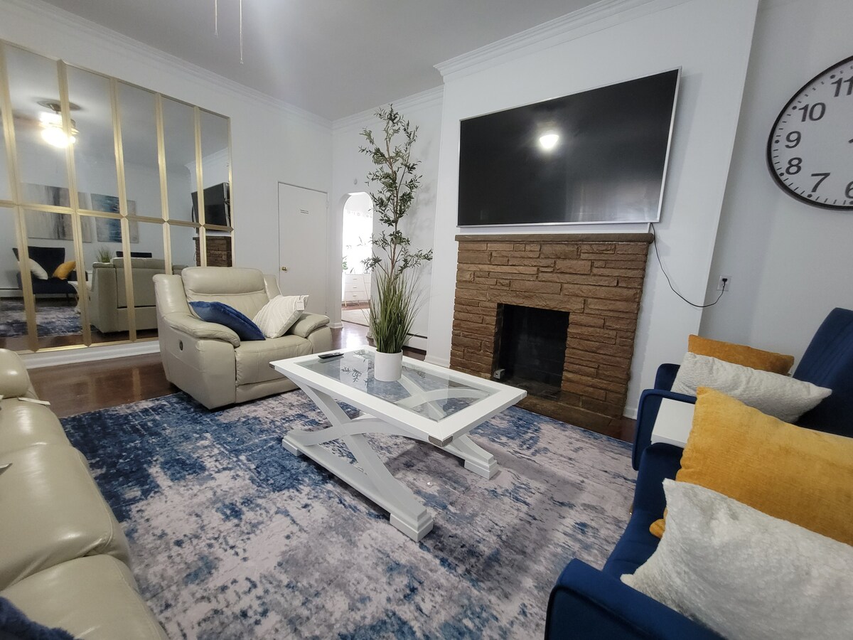 A spacious living area features a light-colored sofa and armchair arranged around a glass-top coffee table. A large television is mounted above a stone fireplace. A decorative plant adds a touch of greenery, and the room is brightened by natural light reflecting off the mirrored wall.