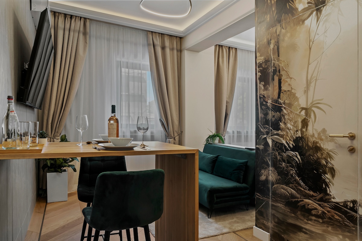 A modern living area features a stylish green sofa adjacent to a decorative wall depicting a lush forest scene. A wooden dining table sets an inviting atmosphere, complemented by elegant glassware and a bottle. Large windows showcase soft drapery, allowing natural light to fill the space.