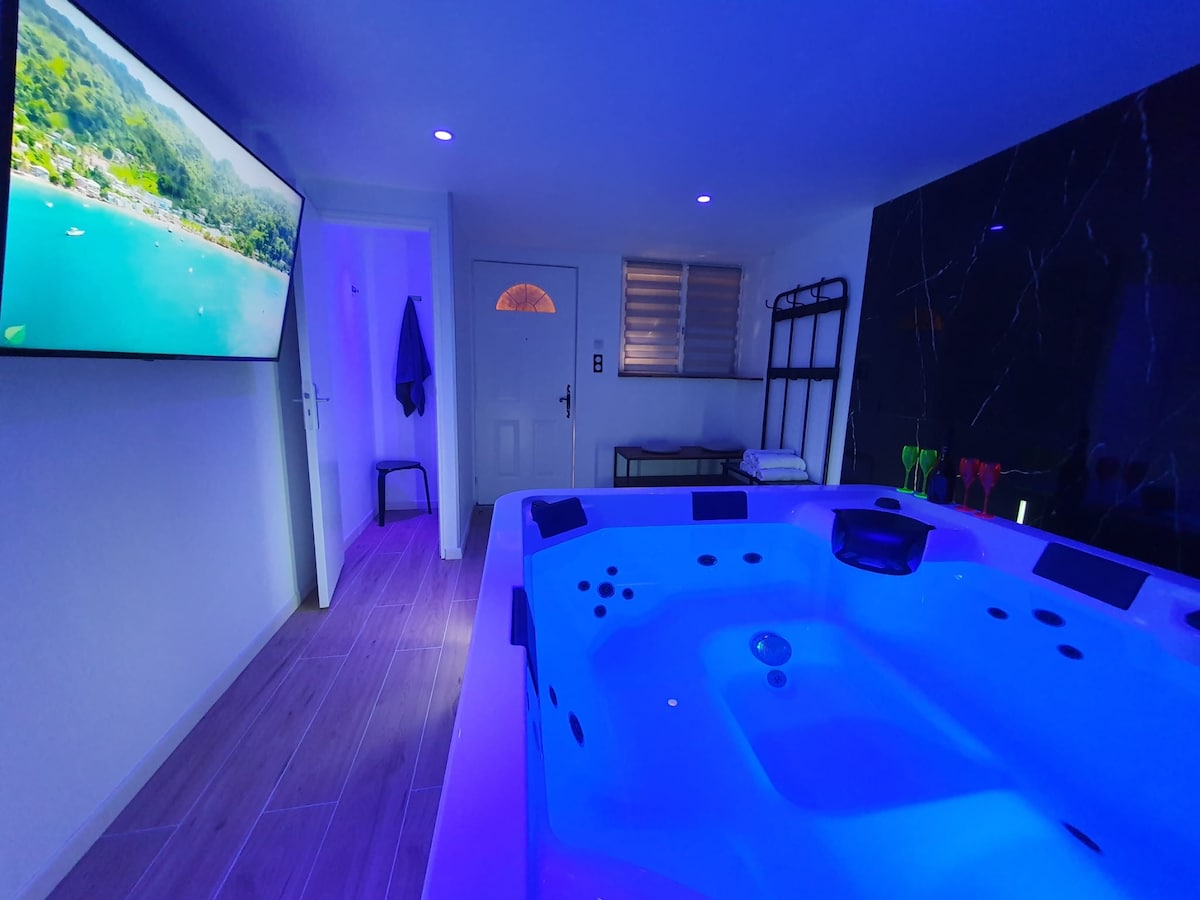 A private spa area is illuminated with soft blue lighting, featuring a spacious jacuzzi with seating for five. A large screen displays a scenic view while a door leads outside. Wall-mounted racks provide storage, enhancing functionality within the serene setting.