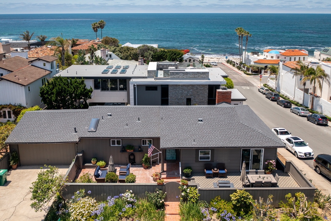 An aerial view of a beach home showcases its proximity to the ocean and street. Lush greenery surrounds the property, with outdoor seating visible on a patio. The ocean is seen in the background, highlighting the home's coastal setting.