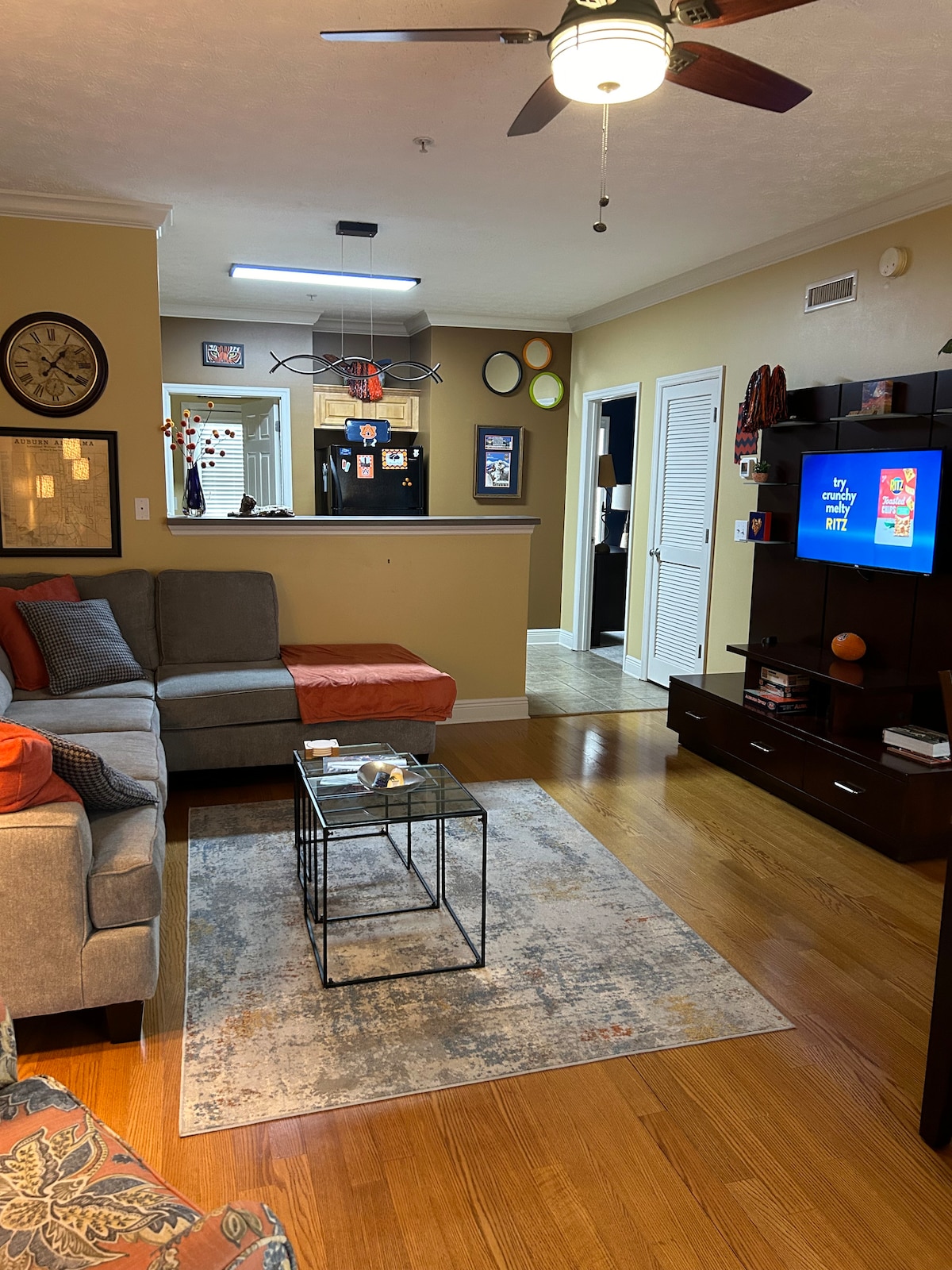 A living area features a spacious sectional sofa with colorful cushions, arranged around a glass coffee table. A flat-screen TV is mounted on a dark wooden unit, while a small kitchen area is visible in the background. Warm lighting enhances the cozy atmosphere.