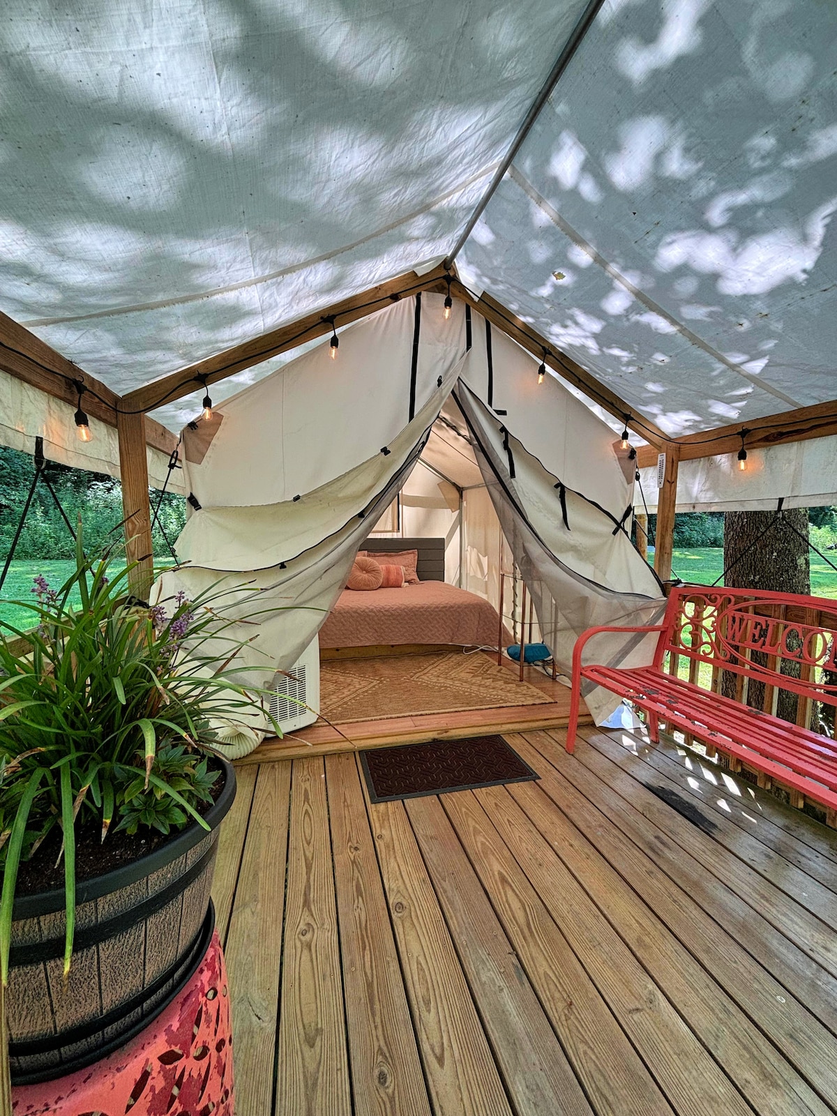 The interior of the cozy glamping tent wagon features a spacious queen bed draped in soft linens. Natural light filters through the zippered openings, illuminating the wooden flooring. An inviting bench sits on the porch, surrounded by potted greenery, creating a connection to the serene outdoor setting.