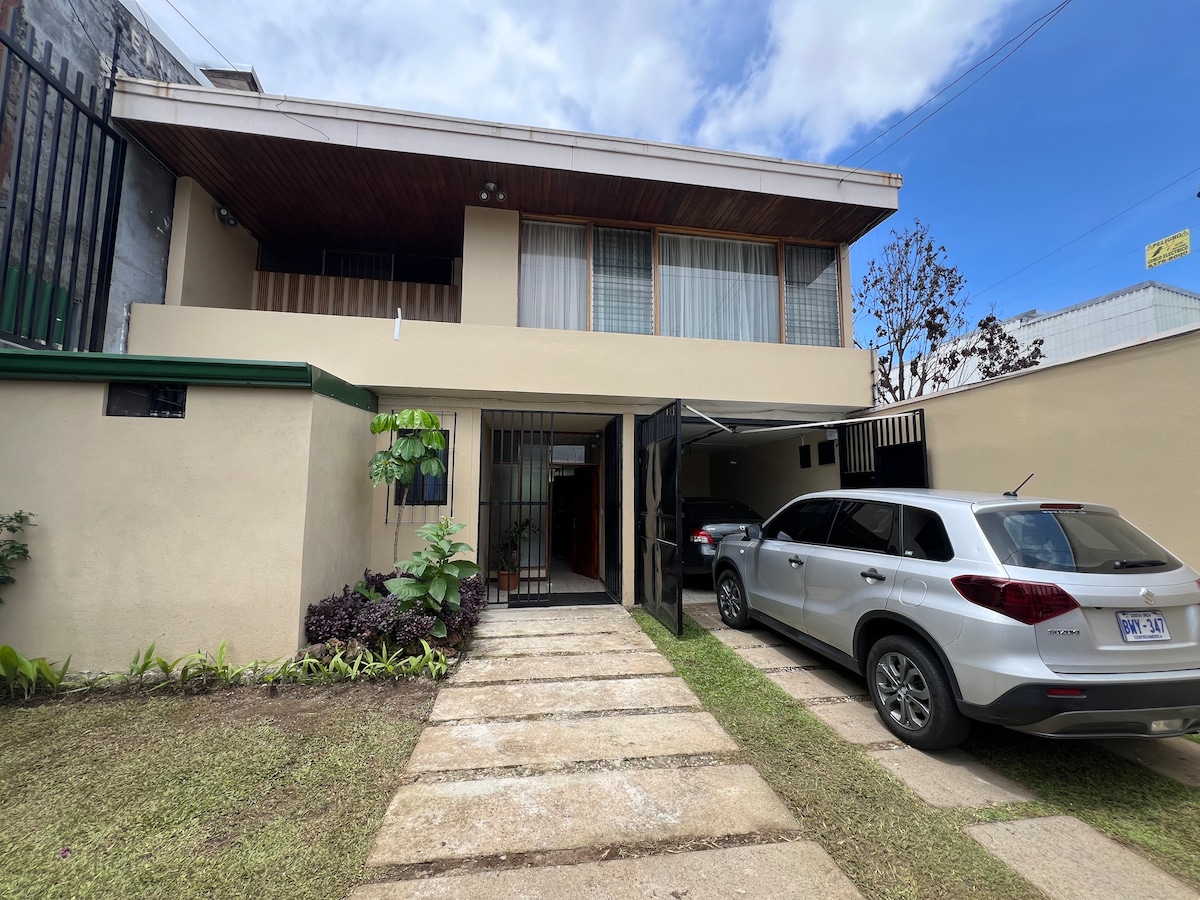 The exterior of a modern home is shown, featuring a welcoming entrance with a pathway leading to the front door. A spacious driveway accommodates two vehicles. Lush greenery is present alongside the pathway, and large windows above suggest ample natural light within.