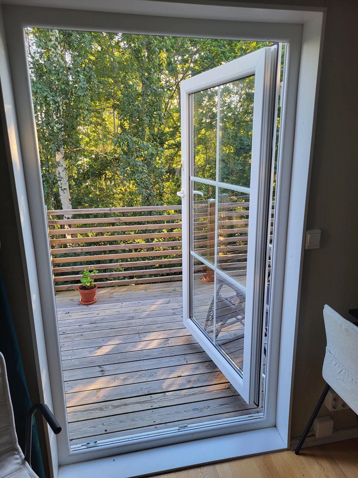 A white door is open to reveal a wooden deck surrounded by greenery. Sunlight casts soft shadows on the deck, while a potted plant adds a touch of nature. The surrounding trees provide a sense of privacy and tranquility.