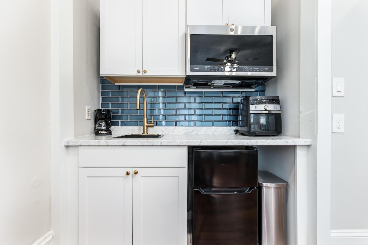 The kitchenette features modern cabinetry in white with gold hardware. A mini fridge and microwave are positioned beneath a chic backsplash. An air fryer sits beside a coffee maker, and a stainless steel trash can completes the functional space, offering essential amenities for guest convenience.