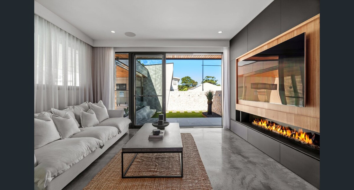 A modern living area features a spacious sofa adorned with soft, neutral cushions facing a sleek coffee table. A contemporary fireplace is set into the wall, with flickering flames visible. Floor-to-ceiling windows allow natural light and provide a view of the outdoor space.