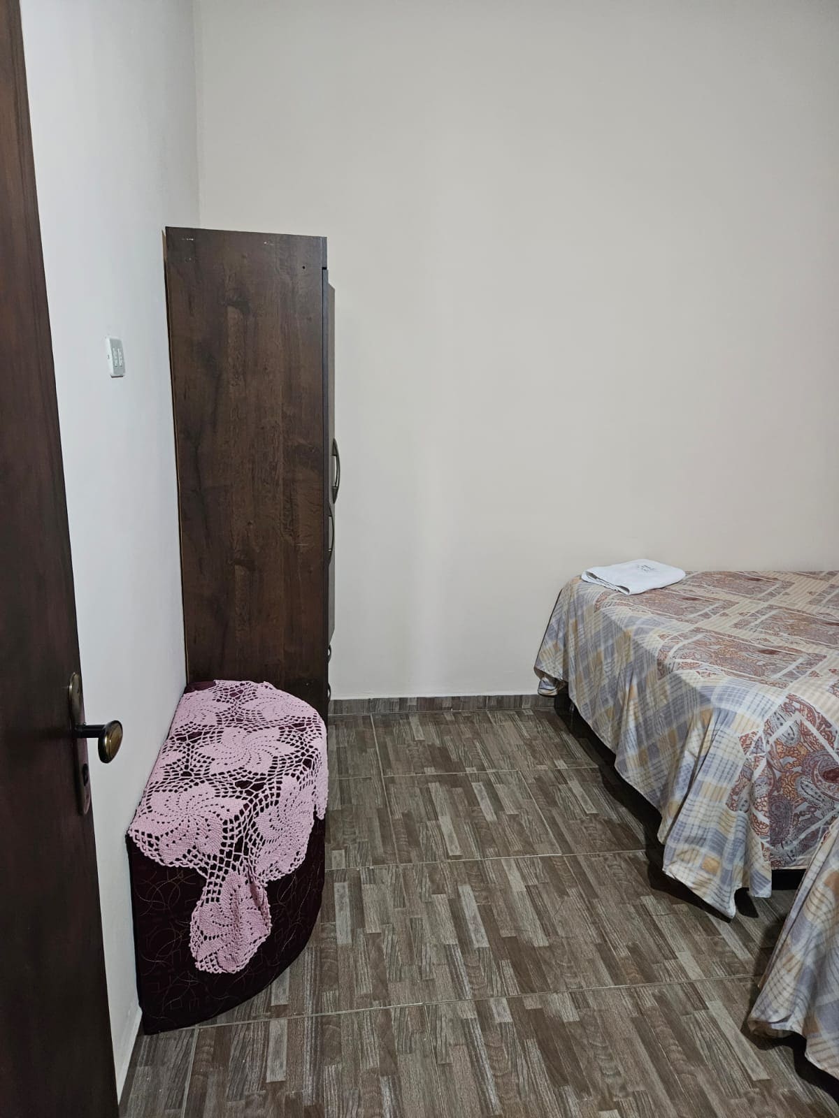 A simple bedroom features a neatly made bed with a patterned coverlet. A wooden wardrobe stands against one wall, while a round Ottoman covered in a lace design is positioned near the bed. The flooring consists of uniform tiles, contributing to a clean and minimalist aesthetic.