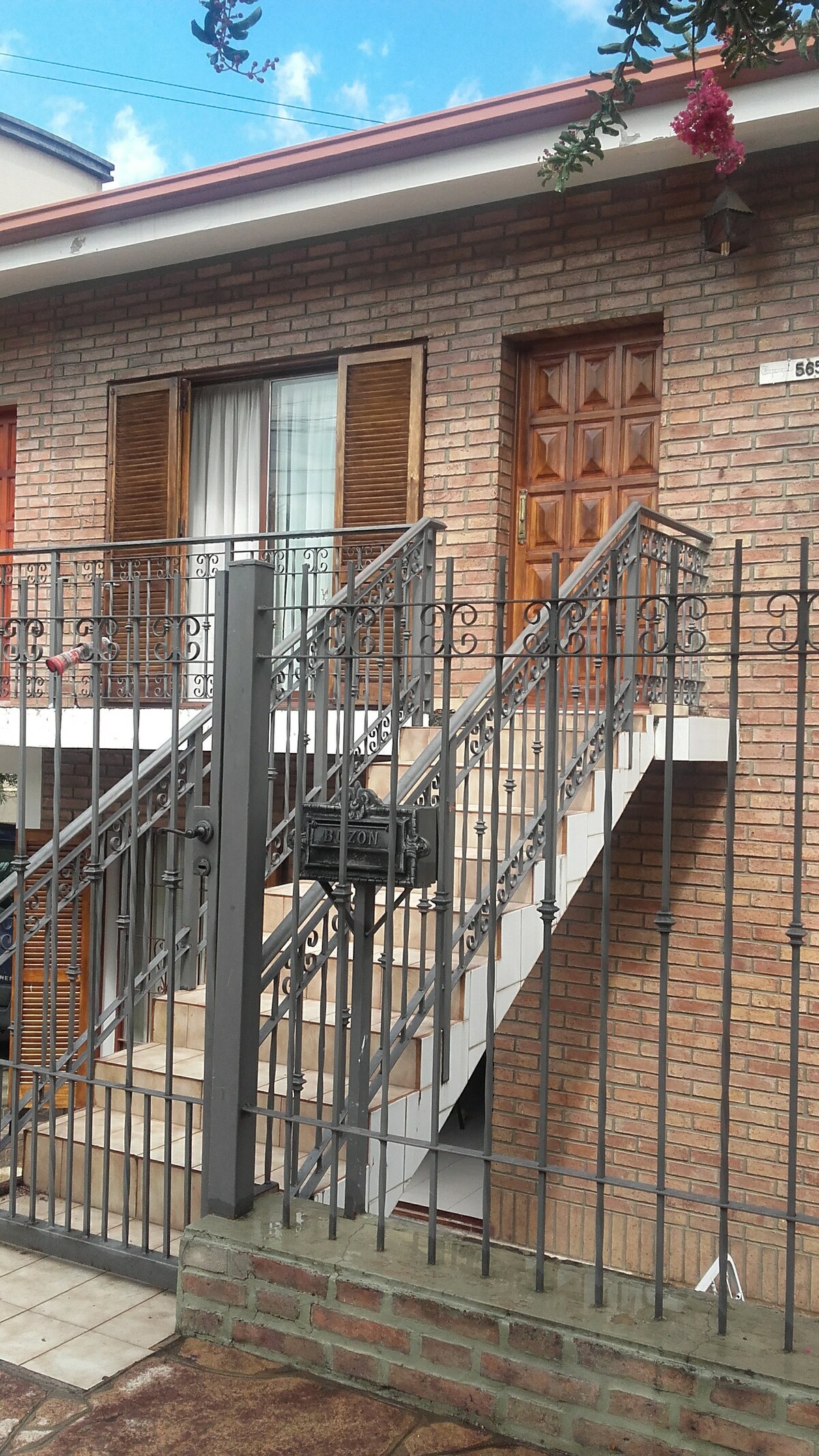 The exterior showcases a brick building with a prominent wooden door and sliding windows adorned by wooden shutters. An iron gate leads to a set of stairs, providing access to the upper level where the apartment is located.