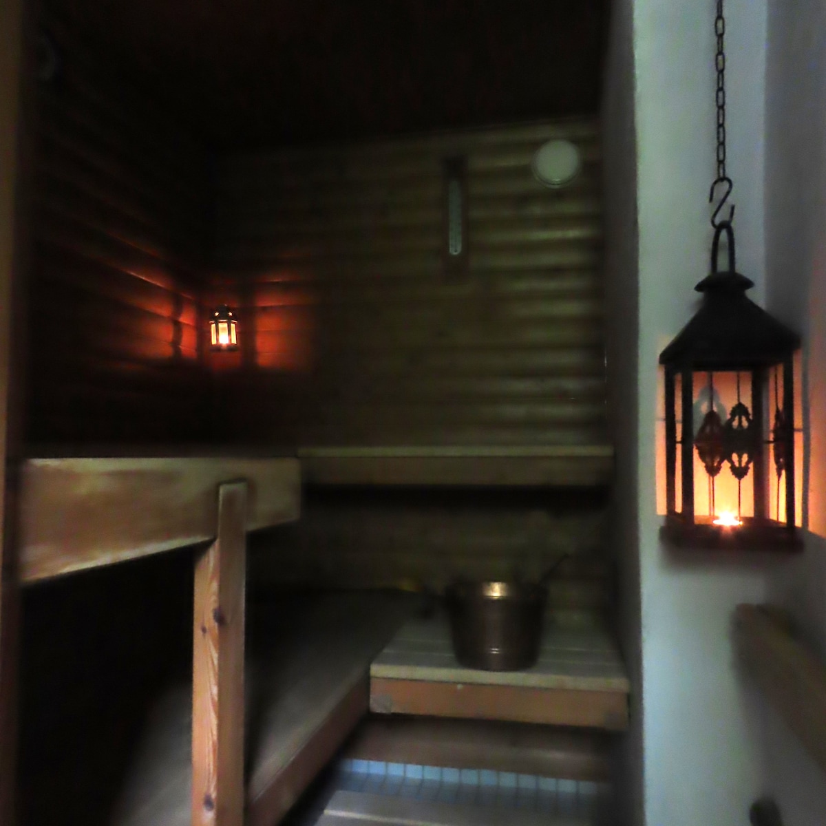 A traditional sauna environment is depicted, featuring wooden benches and warm ambient lighting from lanterns. A copper bucket is placed on a stone floor, complementing the natural wood walls that create a calming space for relaxation.