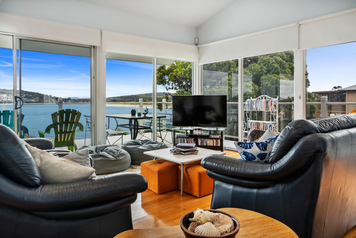 A bright living space features large glass windows providing views of the waterfront and surrounding landscape. Comfortable seating arrangements include leather armchairs and a spacious couch. A flat-screen television is mounted in front of a coffee table, with decorative items placed on it.