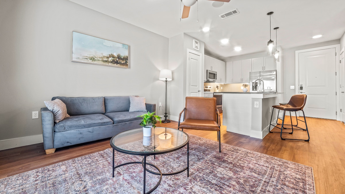 A spacious living area is featured with a blue sofa, two brown chairs, and a round coffee table. A modern kitchen is visible in the background, equipped with stainless steel appliances, and a dining area with a stylish chair and desk. Soft lighting creates a warm ambiance.