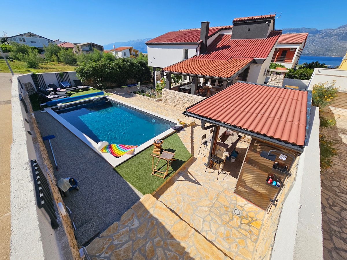 An outdoor area features a swimming pool surrounded by lounge chairs and artificial grass. A stone patio includes a covered terrace with seating and a grill. Scenic views of the mountains and coastline can be seen in the background, enhancing the inviting outdoor setting.