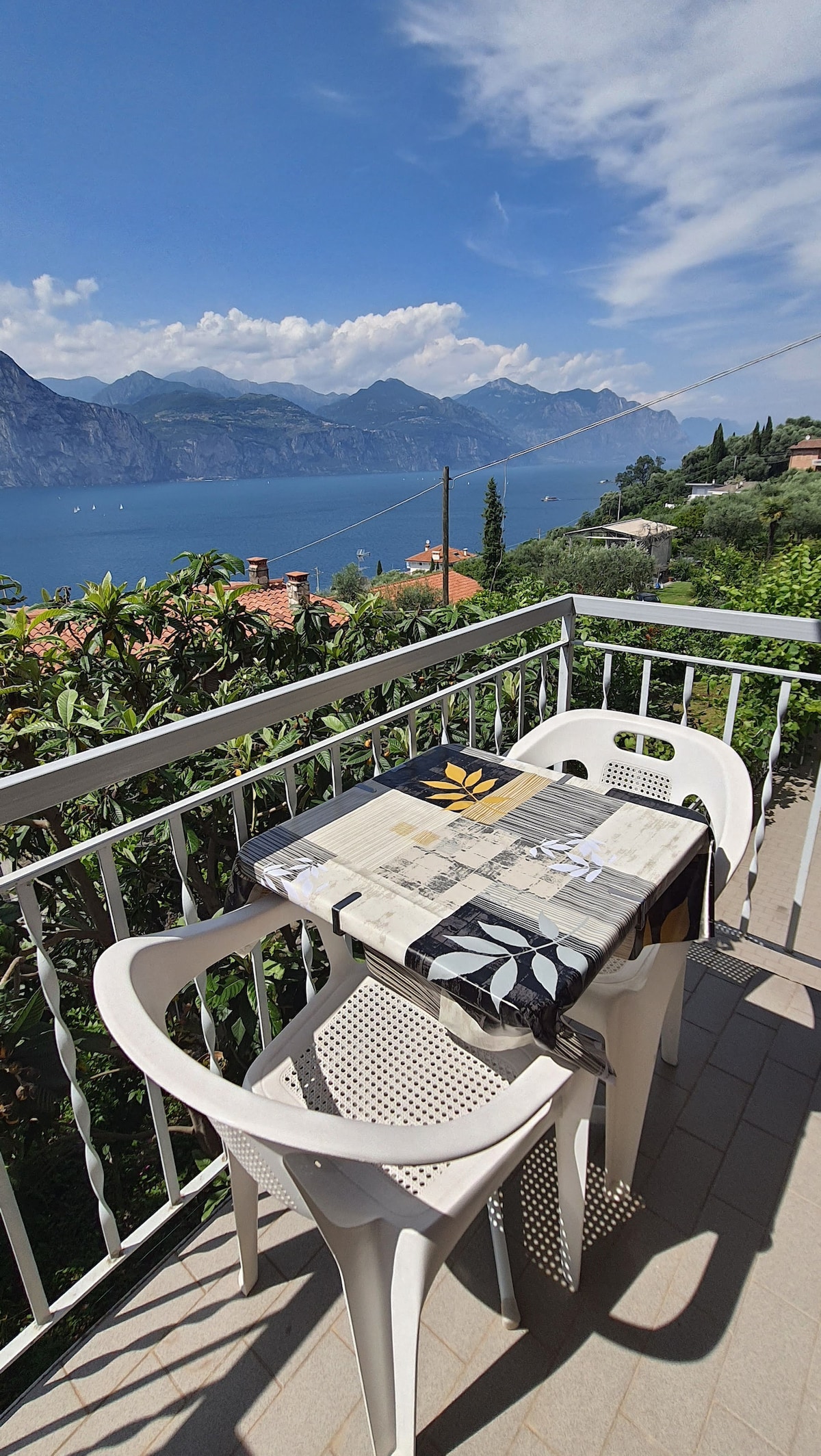A balcony table is set with a decorative tablecloth and surrounded by white plastic chairs. The view of the lake and mountains creates a scenic backdrop, featuring vibrant greenery and distant houses under a partly cloudy sky.