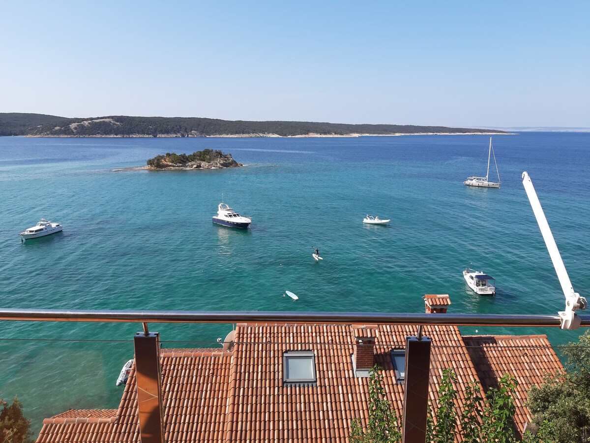 A serene view overlooks a calm sea, with several boats anchored and gently bobbing in the water. The foreground features terracotta roofs, while the horizon presents an unbroken line of the distant island and land, complementing the clear blue sky.