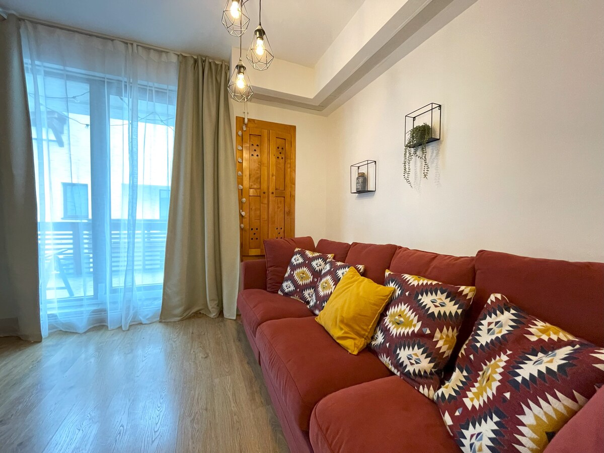 A spacious living area features a soft red sofa adorned with patterned cushions and a bright yellow pillow. Natural light filters through sheer curtains covering a large window, enhancing the warm atmosphere. Decorative wall shelves add a touch of greenery to the neutral walls.