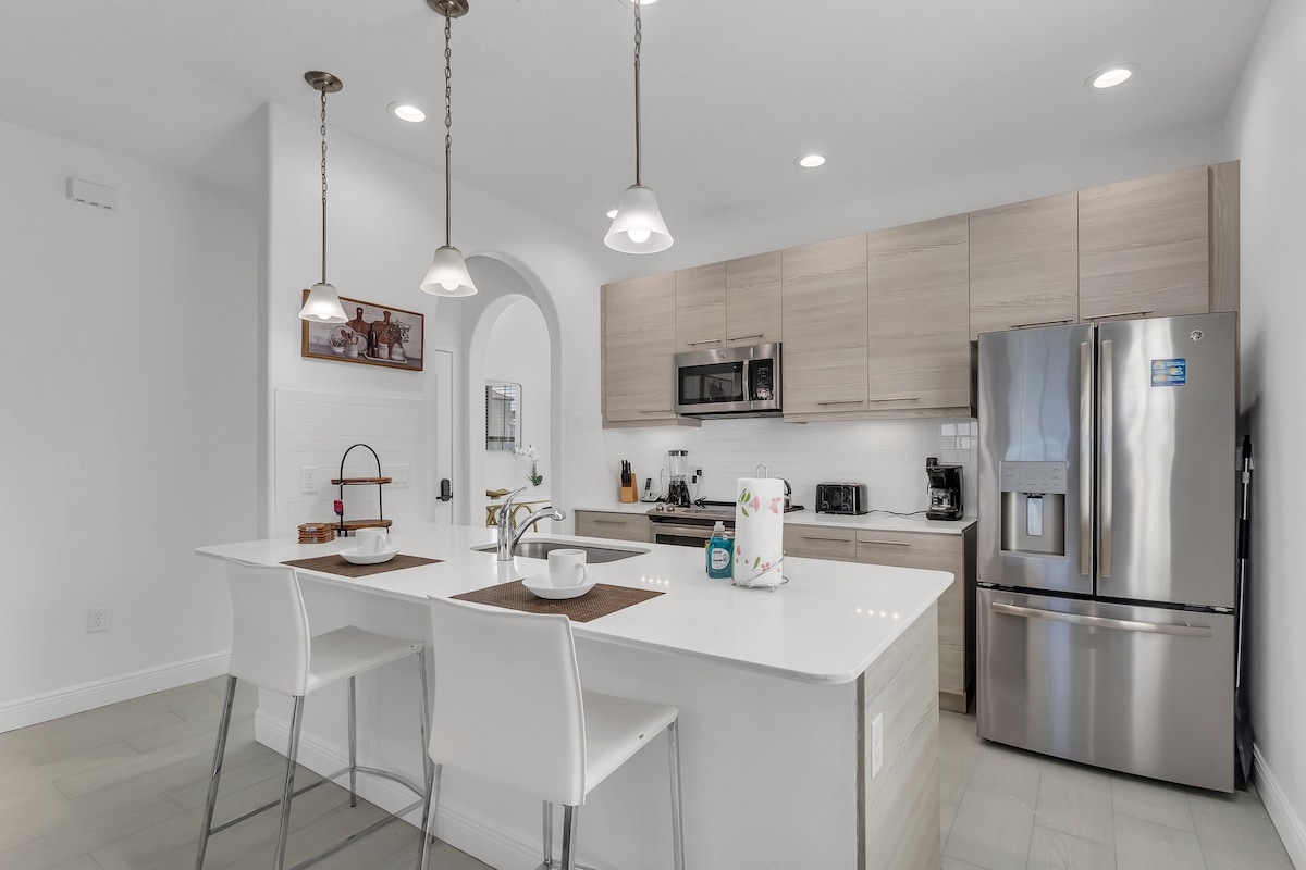 A modern kitchen is equipped with stainless steel appliances, including a refrigerator and microwave. Light wood cabinetry provides ample storage, while a white countertop accommodates two sleek bar stools. Pendant lights hang above, illuminating the space, which features a neutral color palette.