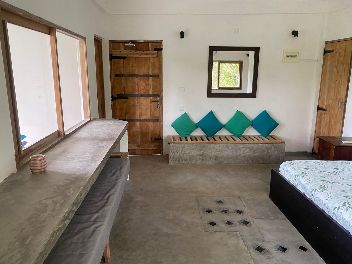 A well-designed apartment interior features a spacious sleeping area with a bed adorned with light bedding. A wooden door and window frames allow natural light to enter. A concrete bench with decorative cushions and a minimalist countertop provide functional space. Neutral tones create a calming atmosphere.