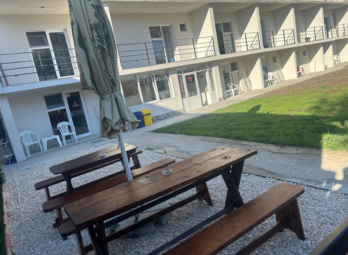 A spacious outdoor area is shown, featuring several wooden picnic tables surrounded by gravel and grass. The building's facade is visible in the background, showcasing a series of rooms with glass doors and railings. An umbrella stands beside the tables, offering shade.