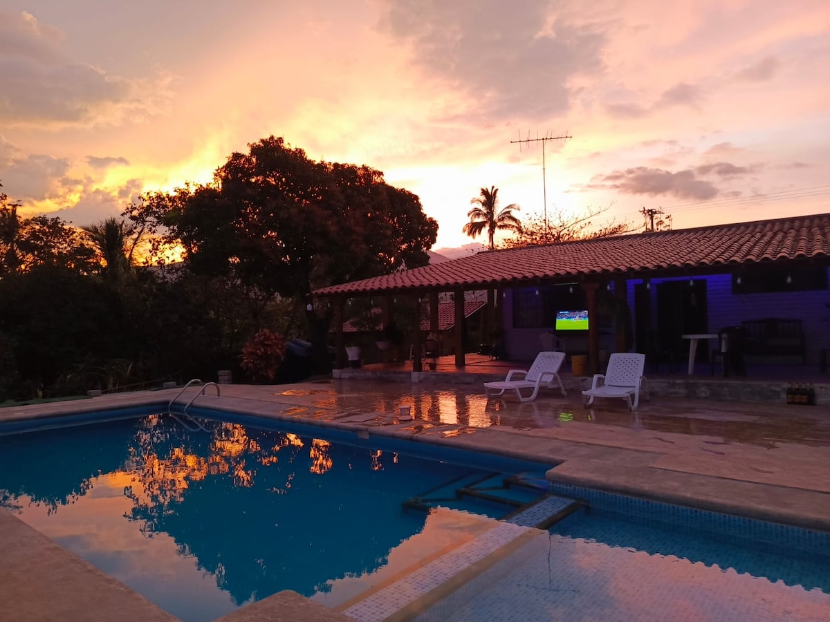 A tranquil swimming pool reflects the warm hues of a sunset, framed by lush greenery and a covered outdoor area. Lounge chairs are positioned beside the water, and the distant structure features a traditional tiled roof.