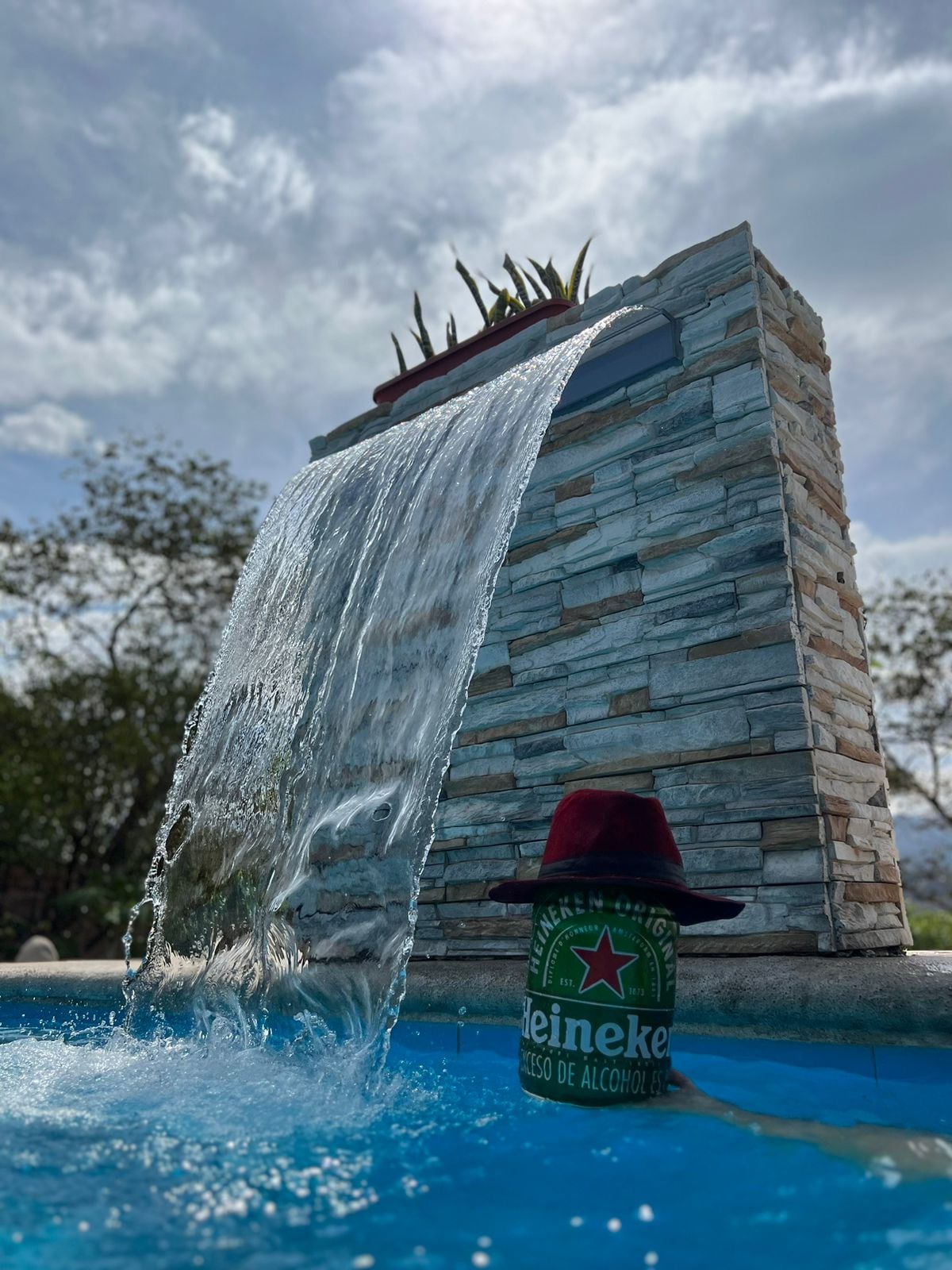 A serene pool scene is captured, featuring a stone waterfall cascading gently into the turquoise water. A green beer can, adorned with a red hat, rests on the pool's edge, surrounded by lush greenery and a clear sky.