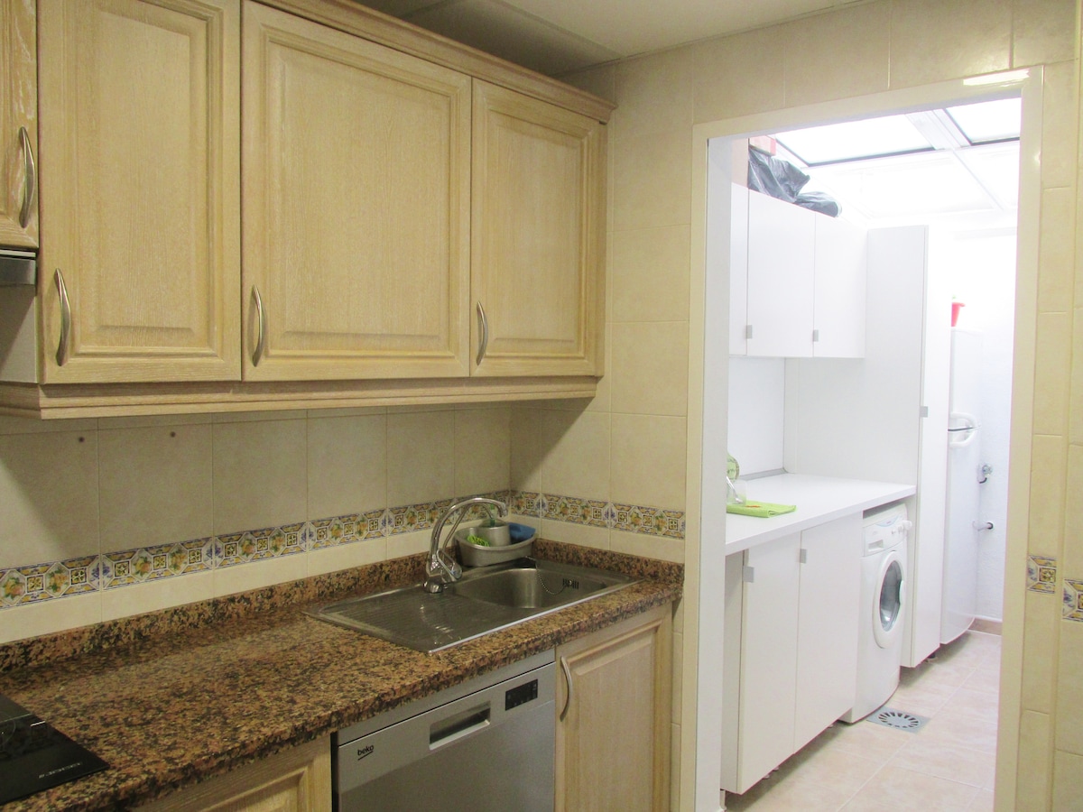 A functional kitchen is equipped with wooden cabinets and a granite countertop. A sink and stainless steel dishwasher are visible, alongside a glimpse of a laundry area featuring a washing machine and a countertop. Clean, neutral tiles adorn the walls and floor.