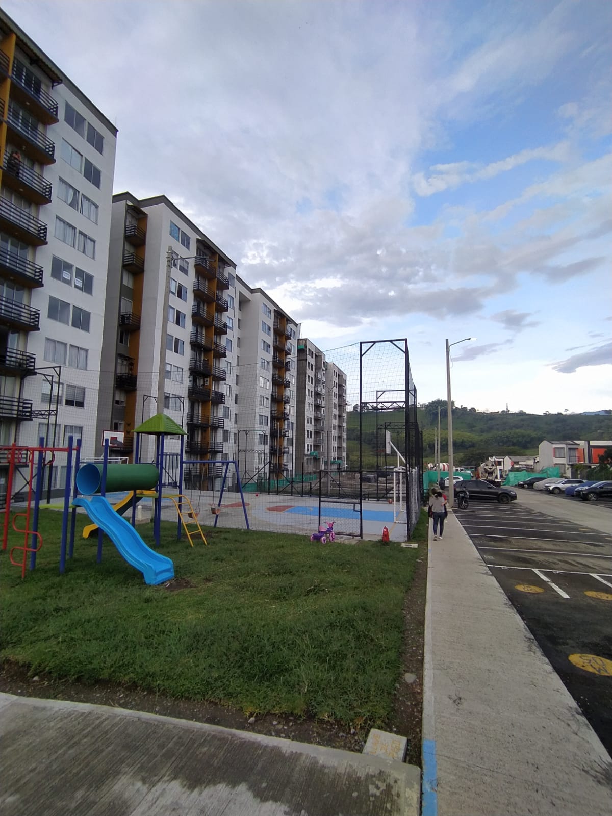 A residential area is visible, featuring multiple apartment buildings with balconies. A playground with colorful structures, including a slide and swings, is situated nearby. A basketball court is present, surrounded by a safety net. Green grass adds a touch of nature to the surroundings.