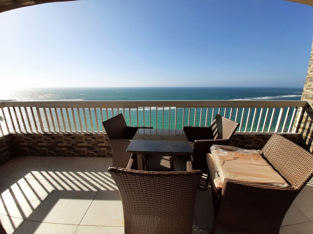 A balcony with stunning ocean views features a glass table surrounded by four wicker chairs. Sunlight casts shadows from the railing, accentuating the serene outdoor space along the water.