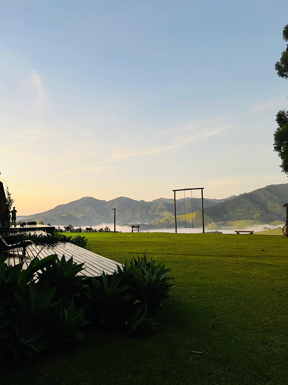 A serene outdoor space features a lush green lawn with a swing set and comfortable seating areas. Mountain views provide a stunning backdrop, while the sky transitions from soft blue to hints of orange, suggesting the approach of sunset. Nearby trees add natural charm.