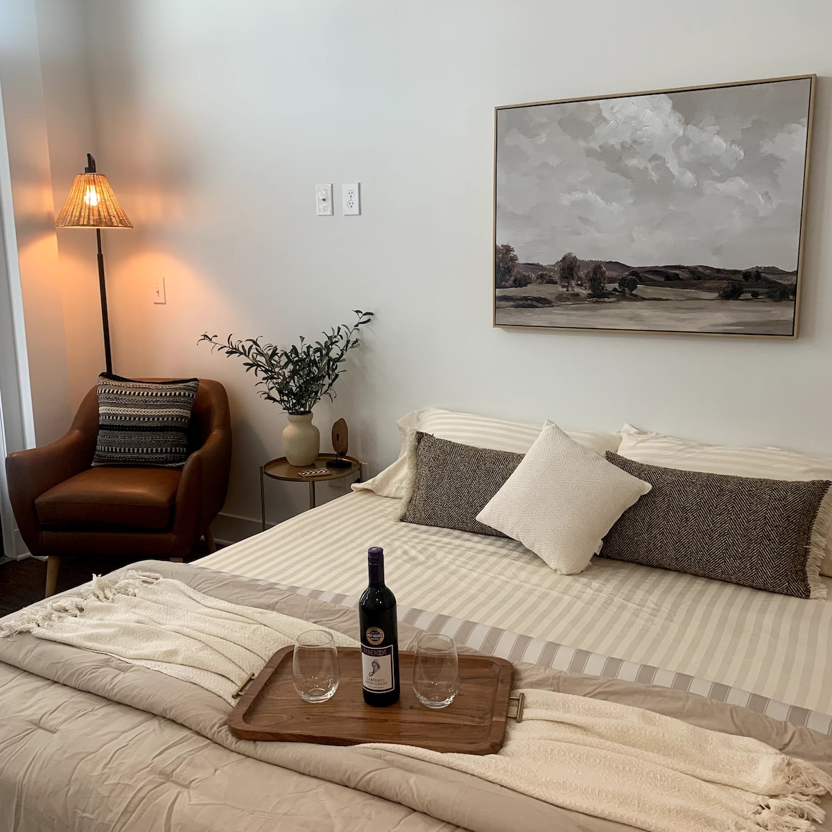 A spacious bed with neutral bedding is featured, complemented by decorative pillows. A small table displays a bottle of wine and two glasses. A comfortable armchair is positioned nearby, with a floor lamp illuminating the corner. A landscape painting is hung above the bed.
