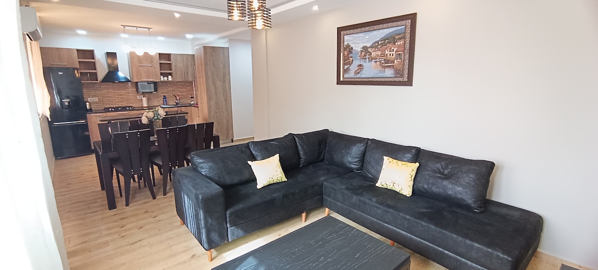 A modern living area features a dark sectional sofa with decorative cushions, facing a coffee table. Behind, a dining table is set for six, and an open kitchen is visible, equipped with various appliances and cabinetry. Light enters through large windows draped with sheer curtains.