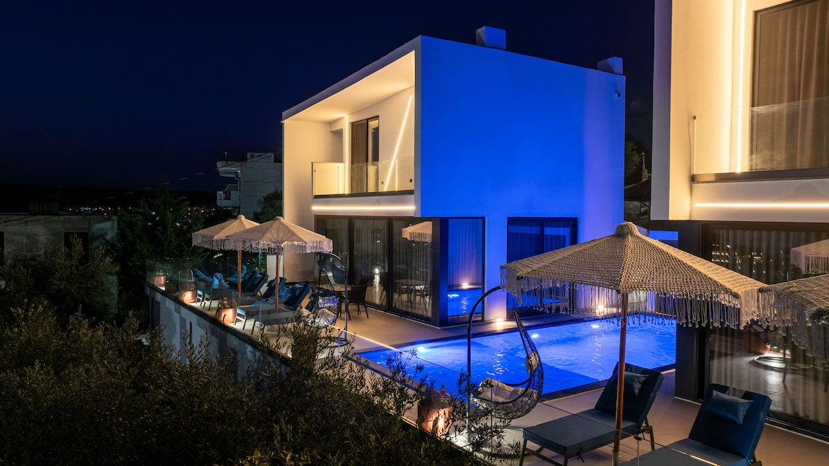 The villa is showcased in evening light, with soft blue illumination highlighting its modern architecture. A shared swimming pool sits in front, surrounded by comfortable lounging areas, including shaded seating under straw umbrellas. Warm lighting enhances the inviting atmosphere of the outdoor space.