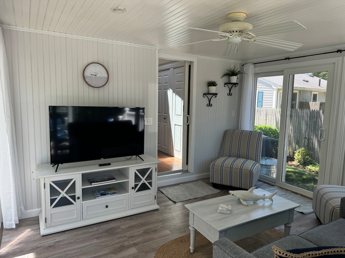 A light-filled living space features a large flat-screen television atop a white media console. Striped accent chairs offer additional seating. A sliding glass door provides access to the outdoor area, while soft natural light filters through sheer curtains, enhancing the inviting atmosphere.