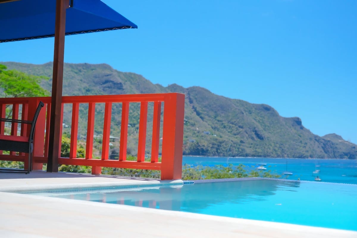 A serene pool area is depicted, featuring a clear blue water surface that reflects the surrounding landscape. A vibrant red railing frames the view, while lush green hills and calm waters are visible in the background under a bright blue sky.