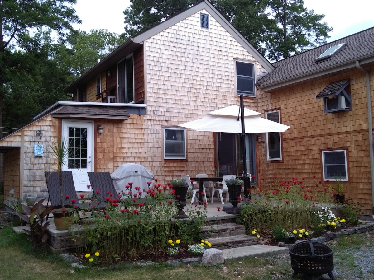 A welcoming exterior features a blend of wooden shingles on the house, showcasing a small patio area with a table and chairs under a white umbrella. Flower beds with vibrant blooms surround the steps, adding color to the outdoor space.