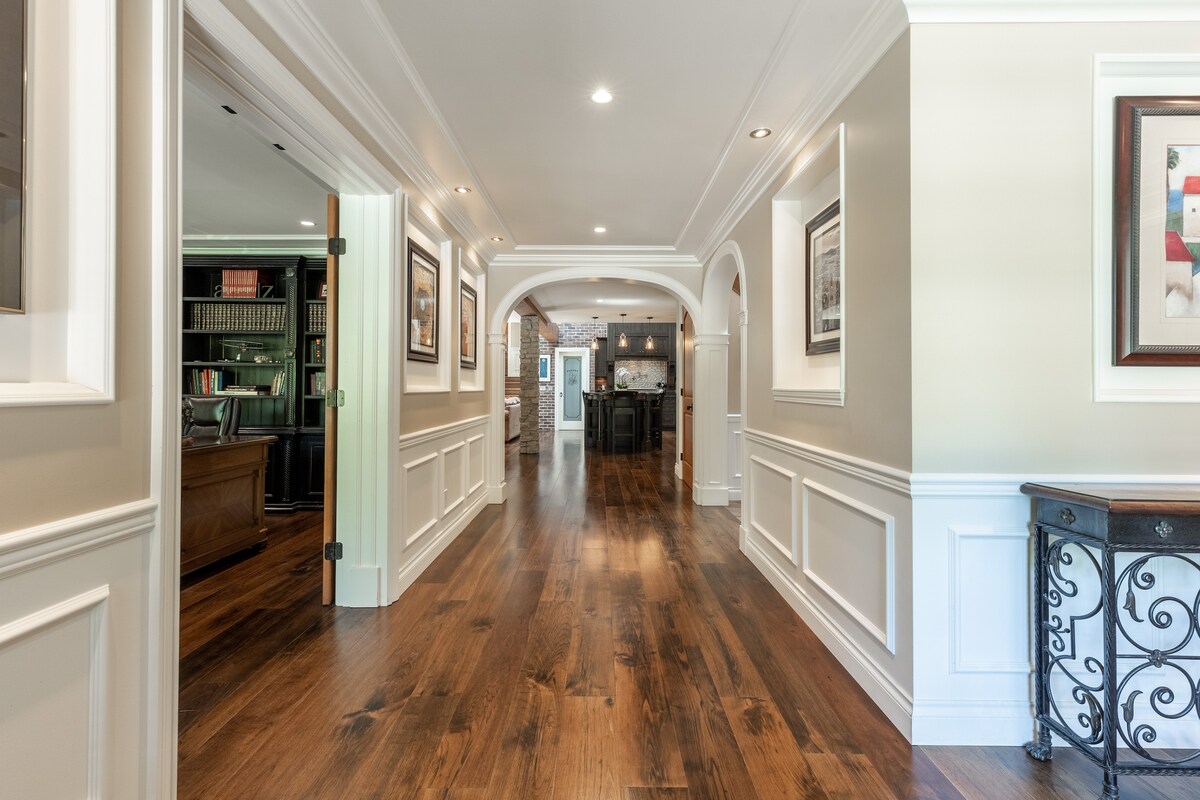 A wide corridor showcases polished wooden flooring and white wainscoting, complemented by arched doorways. The space is illuminated with recessed lighting, while framed artwork adorns the walls, leading towards a doorway that opens into a stylish kitchen area.