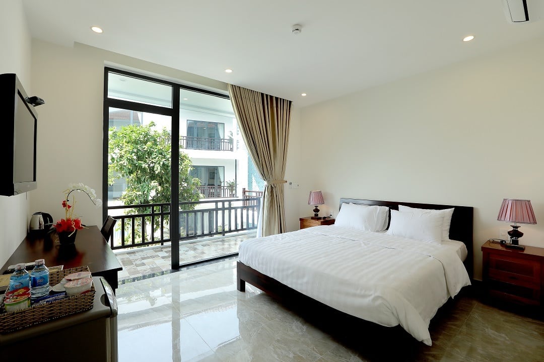 A spacious double room features a king-size bed centered against a light-colored wall. Large glass doors open to a balcony, allowing natural light to fill the space. Complementing the room are bedside tables, a TV, and a small seating area, enhancing comfort.