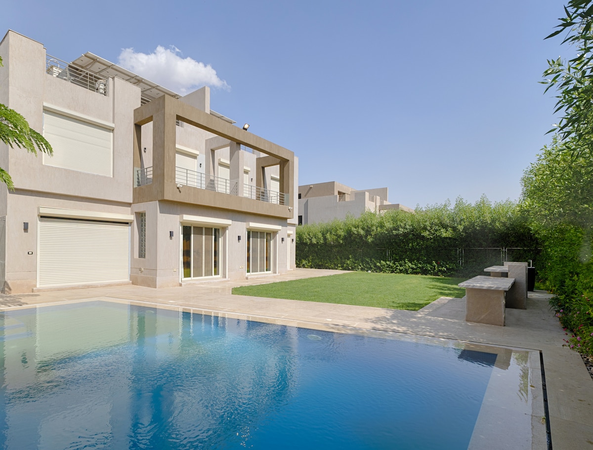 A serene outdoor space features a swimming pool surrounded by a well-maintained garden. The exterior of the property showcases modern architectural elements, including large glass doors that open to the pool area and a spacious stone bench alongside the water.