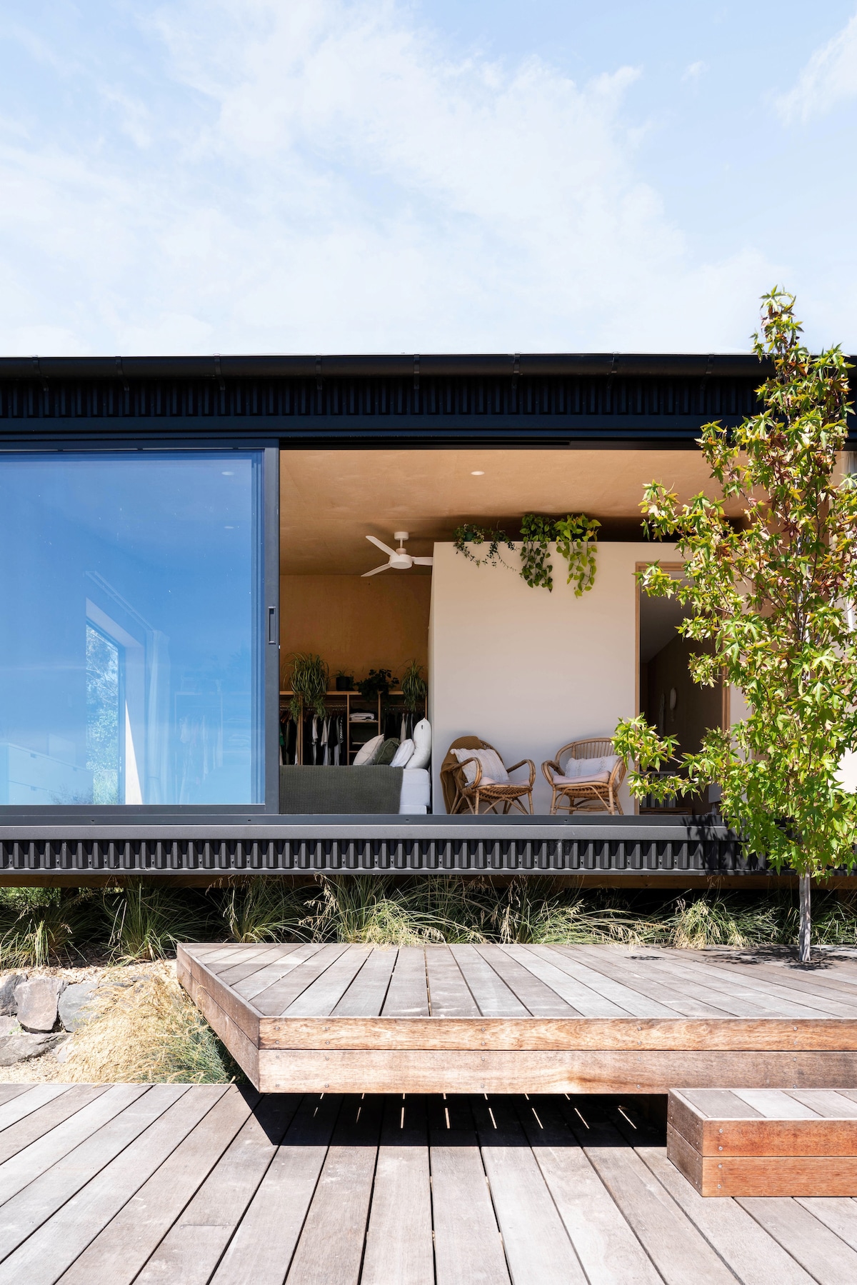 A modern exterior view of the house shows a large glass sliding door opening to an inviting indoor space. Timber decking extends in front, while greenery is visible both inside and outside. A ceiling fan can be seen, enhancing airflow within the stylish area.