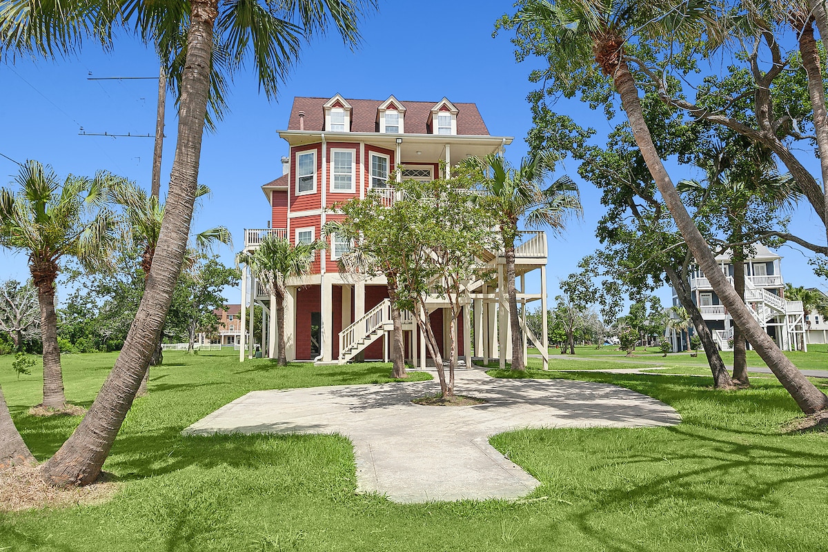 A multi-story house with a vibrant red exterior is surrounded by lush green grass and palm trees. Its elevated structure is supported by a concrete foundation, with a wide staircase leading to the entrance. A clear blue sky serves as a backdrop.