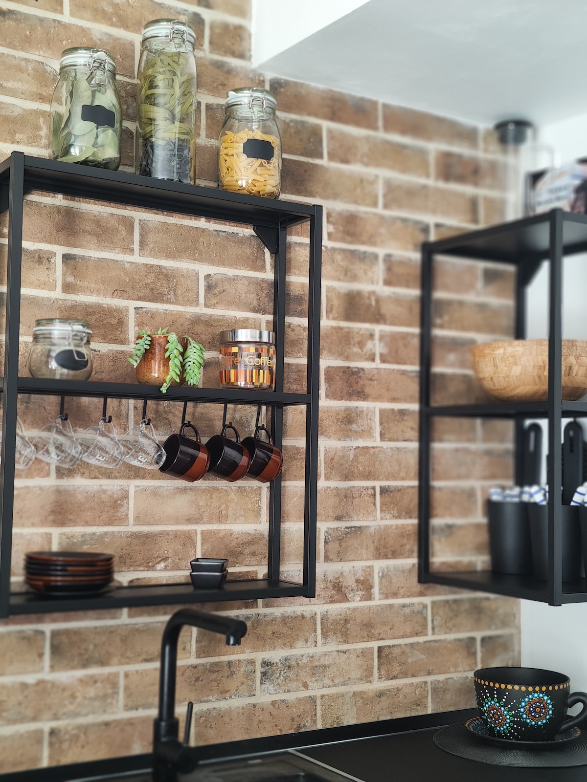 A modern kitchen display features open shelving against a warm, brick wall. Glass jars filled with various ingredients are visible alongside hooks holding several small cups. A dark countertop is complemented by plates and bowls below, enhancing the functional aesthetic of the space.