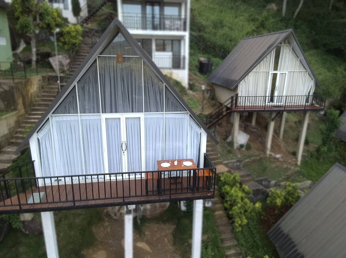 The image shows two triangular-roofed structures elevated on stilts, surrounded by greenery. Each cabin features expansive glass doors, providing a seamless view of the outdoors. A wooden table with two chairs is positioned on the balcony of the nearer cabin.