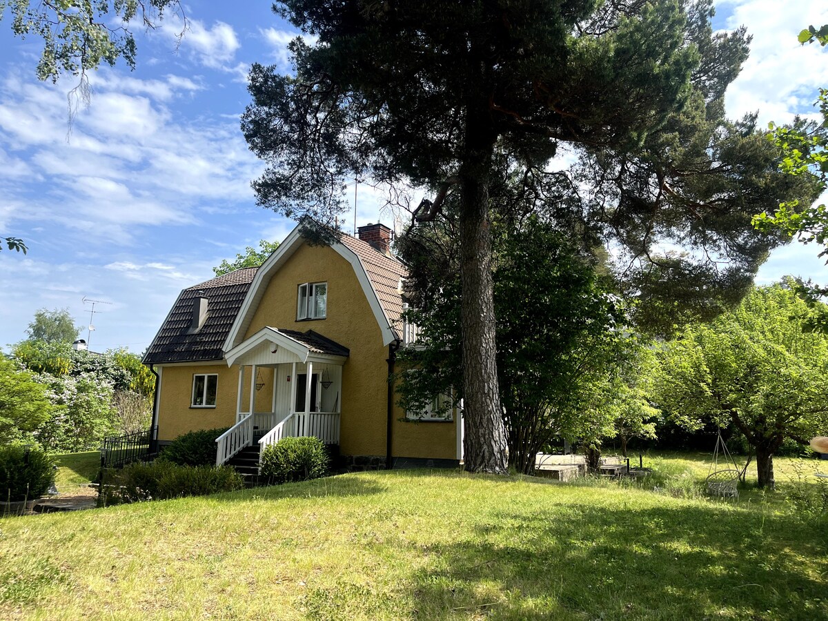 A charming two-story yellow house is set amongst greenery, with a large tree providing shade in the foreground. The inviting entrance features a covered porch and steps leading to the front door. The spacious yard offers room for outdoor activities.
