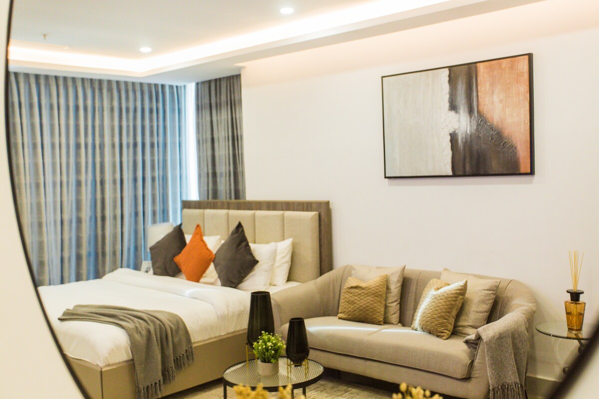 A well-appointed room features a spacious bed dressed in soft linens, complemented by decorative pillows. A cozy seating area with a light-colored sofa and accent pillows is positioned near a round coffee table. Large windows with striped curtains provide a softly lit ambiance.