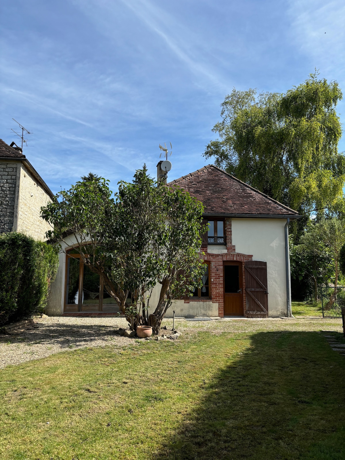 The exterior of the house showcases a quaint two-story design with a sloped roof. A large tree is present in the yard, surrounding a well-kept garden area. Sunny skies provide bright natural light, enhancing the tranquil setting provided by the greenery.