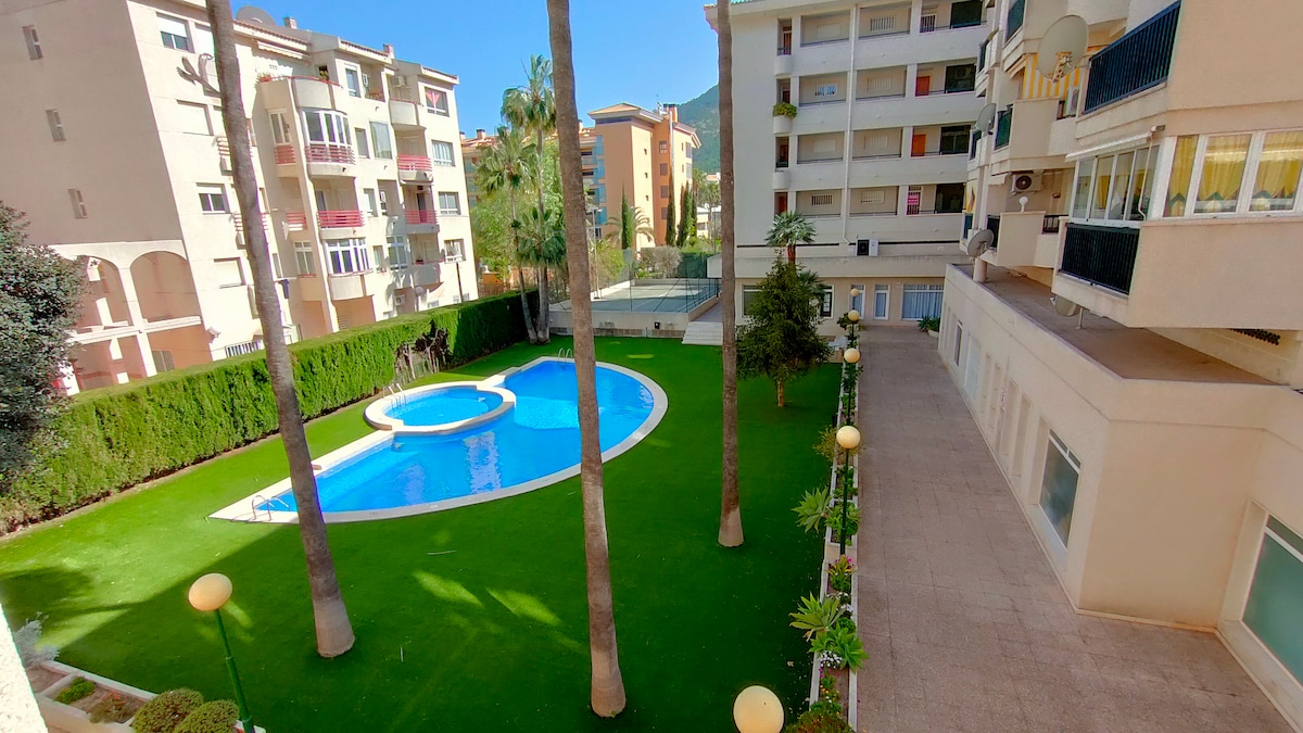 A well-maintained outdoor area is presented, featuring a sparkling pool surrounded by green grass and palm trees. The space includes neatly arranged garden plants and patio seating. Residential buildings frame the scene, creating a serene environment for relaxation.