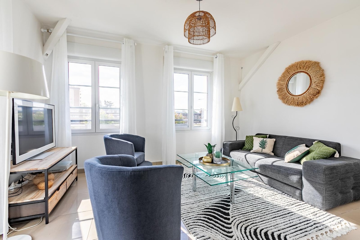 A light-filled living area features large windows framed by white curtains, which enhances the bright ambiance. A comfortable gray sofa is accompanied by two blue armchairs. A glass coffee table rests on a patterned rug, with a decorative plant positioned on top.
