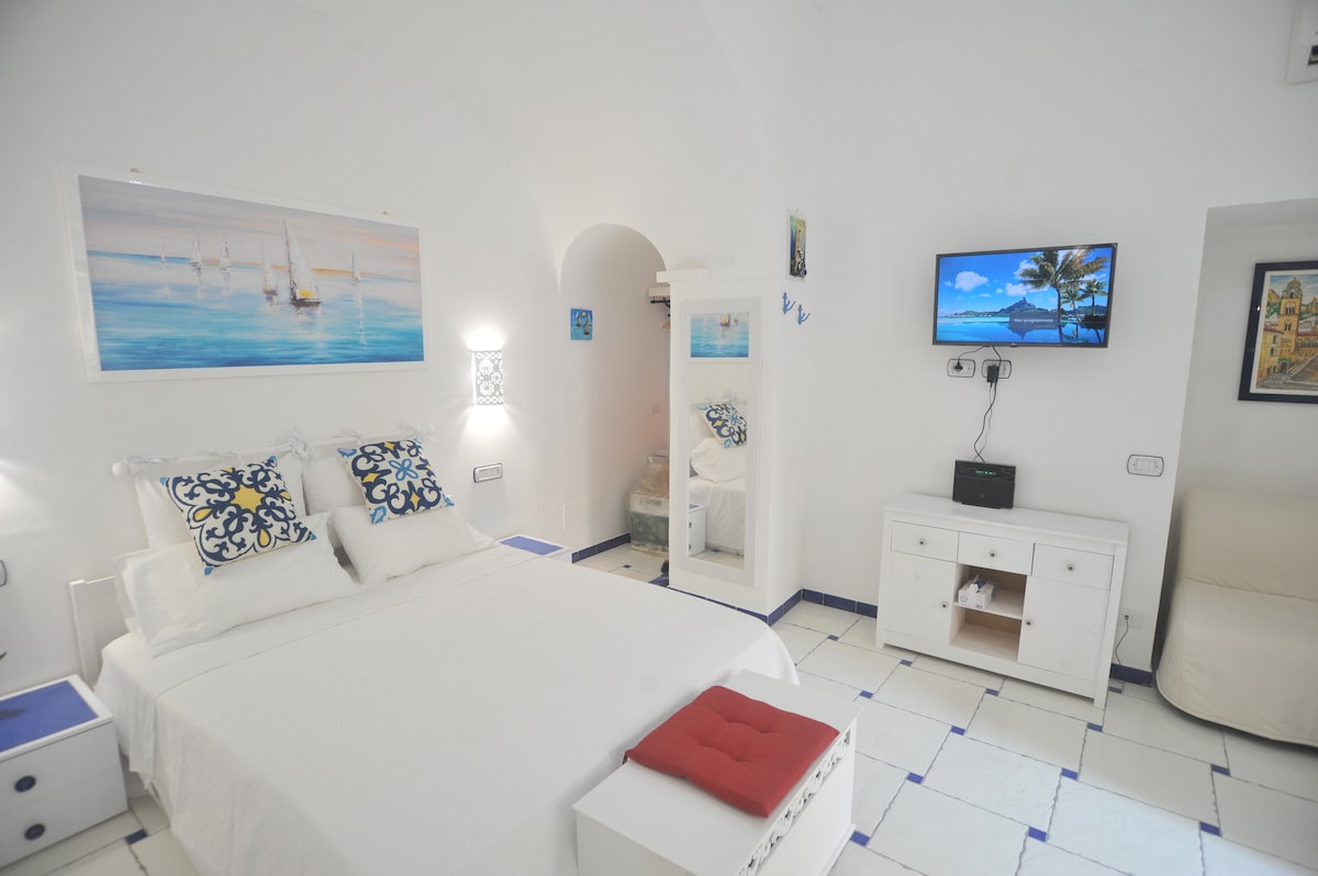 A bright bedroom is presented with a spacious bed adorned with decorative pillows. A wall-mounted television is visible, along with a mirror and a cozy seating area. The light-colored walls and tiled flooring contribute to a serene ambiance.