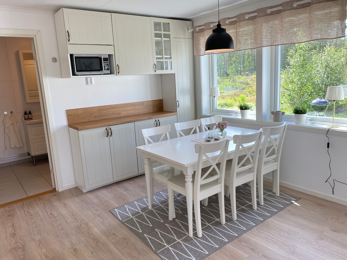A bright dining area features a white table surrounded by six chairs, positioned on a light grey area rug. Soft natural light filters through large windows, enhancing the light cabinetry and wooden countertop. Small decorative items and plants add a touch of warmth to the space.
