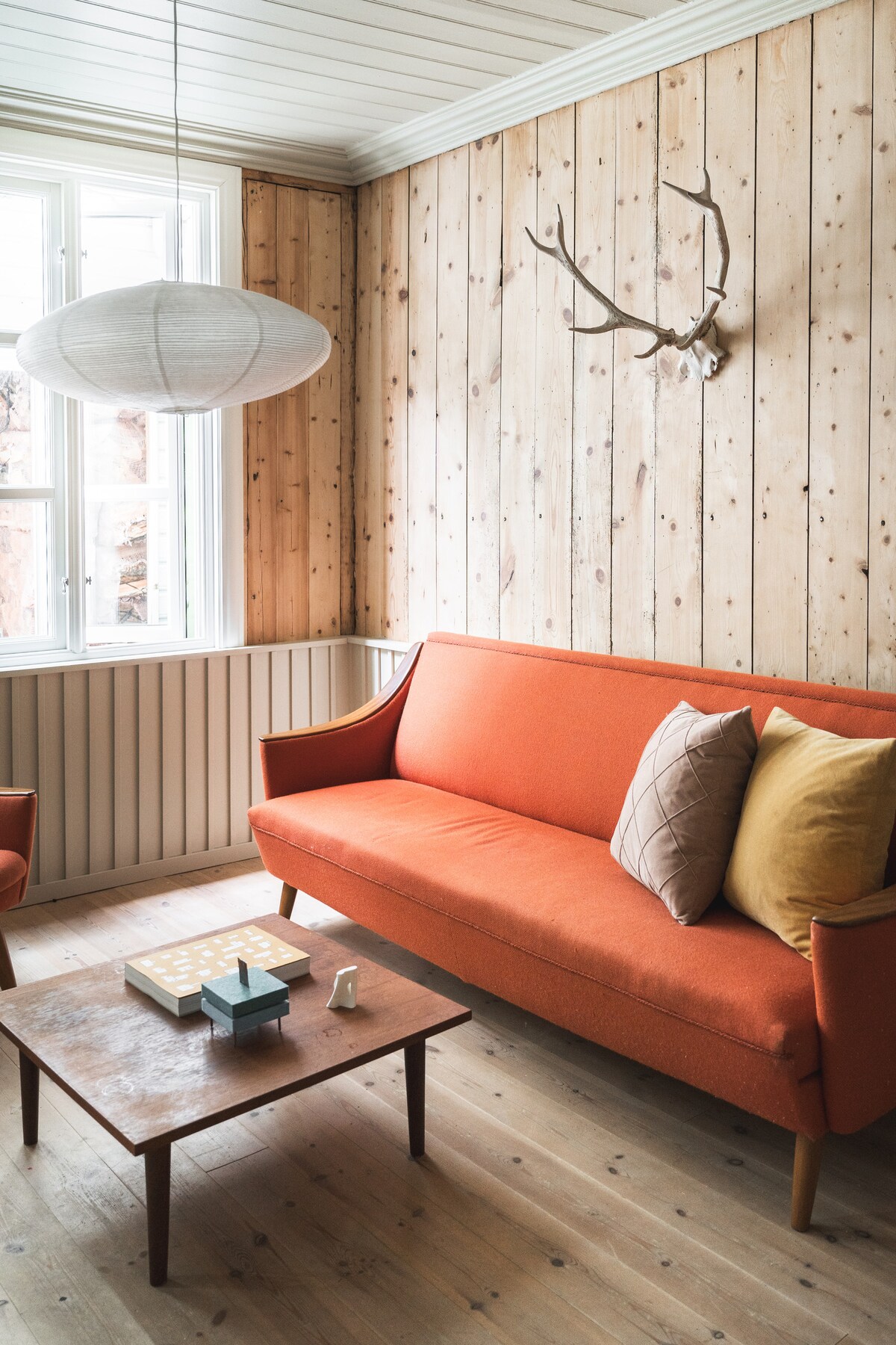 A cozy living area features a bright orange sofa complemented by pillows in earthy tones. A light wood table sits at the center, adorned with simple decor. Natural light enters through a large window, enhancing the warmth of the wooden wall paneling.